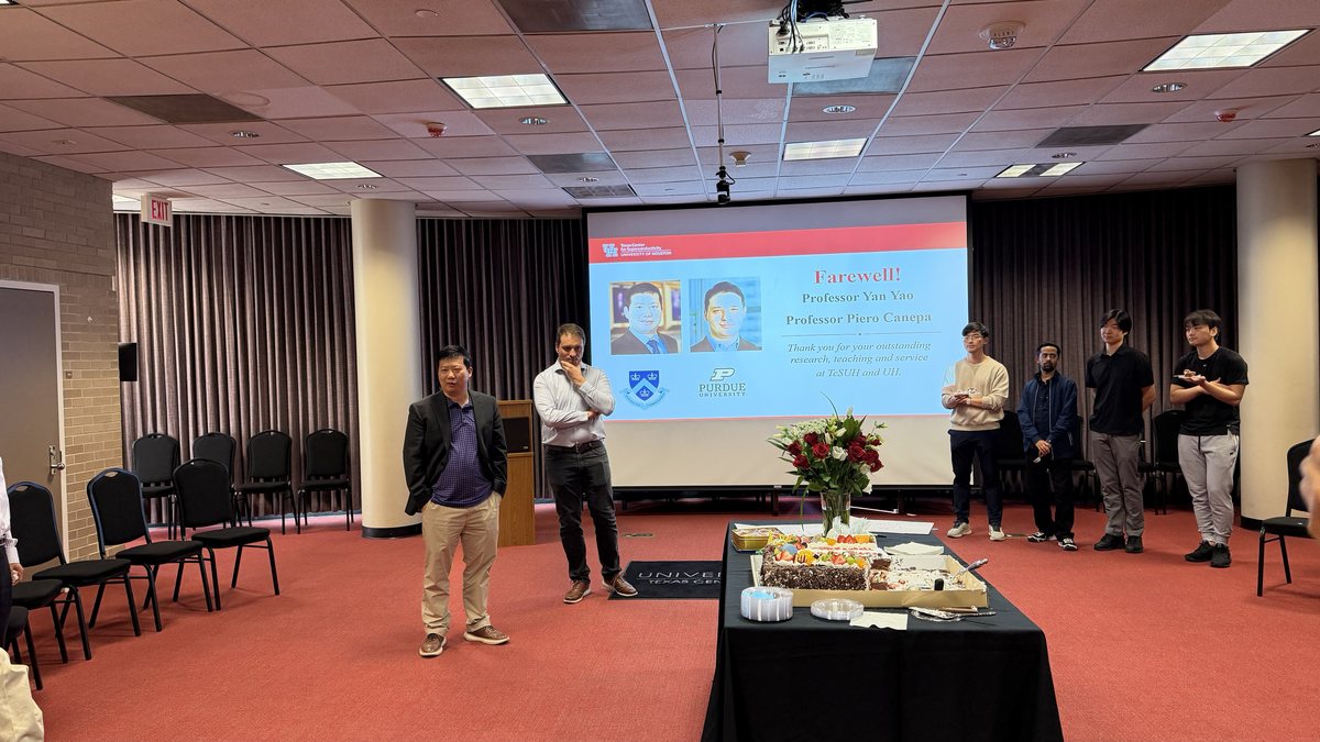 Two men standing before a screen reading "Farewell! Professor Yan Yao, Professor Piero Canepa", beside a table with a cake and flowers, as others look on